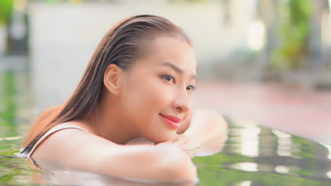 Primer-Plano-De-La-Cara-De-Una-Mujer-Asiática-Con-Una-Piel-Perfecta-Apoyada-En-Los-Brazos-Y-Sonriendo-Al-Borde-De-La-Piscina-En-El-Resort-Tópico-En-Bali