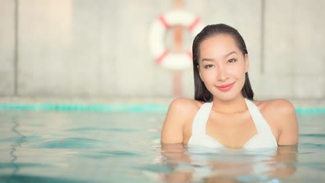 Attractive-cheerful-Thai-young-woman-immersed-in-pool-water-takes-bath-with-lifebuoy-in-background