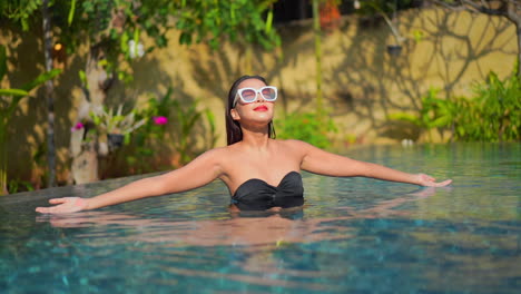 Smiling-happy-Asian-lady-in-a-swimming-pool-wearing-a-black-bikini-and-white-sunglasses-in-Tropical-hotel