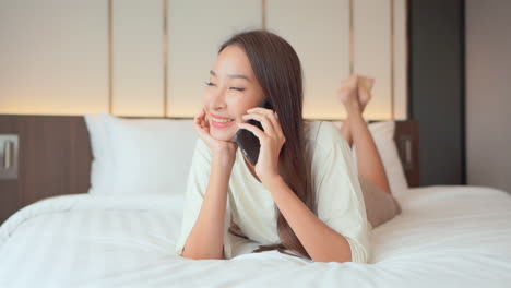 Happy-and-beautiful-Thai-young-woman-lying-on-bed-talks-on-mobile-phone
