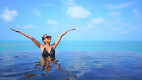 Half-submerged-beautiful-woman-standing-in-the-infinity-pool-rising-her-arms-above-her-head-on-a-blue-sky