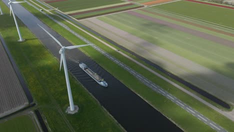 Dutch-rural-infrastructure-with-canal-and-moving-ship,-green-renewable-energy-from-windmill-and-traditional-colorful-tulips,-aerial