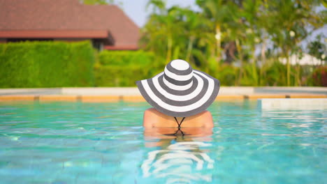 Back-view-of-woman-with-large-striped-hat-bathing-in-luxurious-resort-swimming-pool