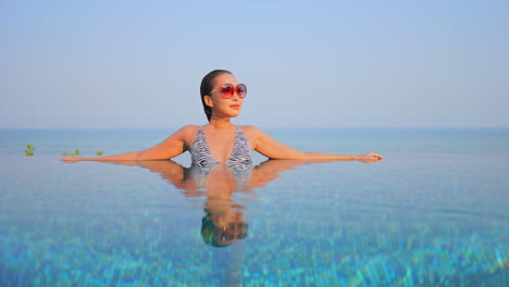 Asian-young-woman-leaning-on-the-border-inside-infinity-pool-front-view-daytime-cloudless-sky,-template-copy-space