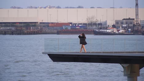 young-female-in-a-distance-is-walking-towards-the-edge-of-a-platform-build-on-water-to-relax-by-the-river-wearing-black-jacket-and-shorts-near-to-docks-industrial-environment-windy-weather-slow-motion