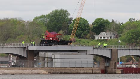 Small-crew-of-men-walking-along-side-a-moving-crane-to-watch-the-dam-rollers-before-lowering-them-into-position-at-Lock-and-Dam-Number-14-on-the-Mississippi-River,-near-LeClair,-Iowa-USA
