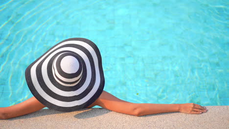 Back-of-a-woman-in-Floppy-stripped-hat-leaning-on-pool-border-inside-swimming-pool,-template-copy-space