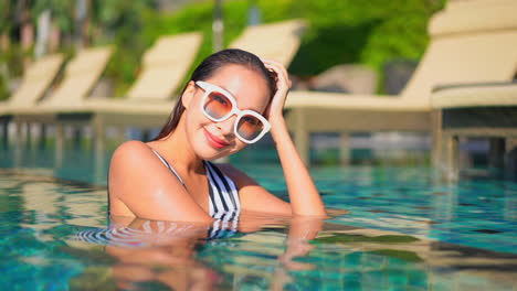 Portrait-of-Asian-woman-inside-the-swimming-pool-water-looking-ar-camera-wearing-white-sunglasses-and-leaning-on-border-next-to-the-line-of-deck-chairs