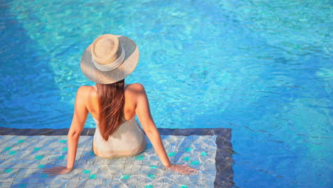 Rückansicht-Der-Fitten-Frau,-Die-Im-Seichten-Wasser-Am-Rand-Eines-Swimmingpools-Sitzt,-Einen-Sonnenhut-Trägt-Und-Sich-Auf-Den-Malediven-Auf-Ihre-Arme-Stützt