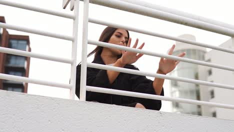 Mysterious-pretty-woman-with-big-brown-eyes-wearing-black-jacket-and-white-t-shirt-is-hiding-behind-white-fence-railing-touching-it-looking-cute-flawless-around-beautiful-houses-in-a-city-slow-motion