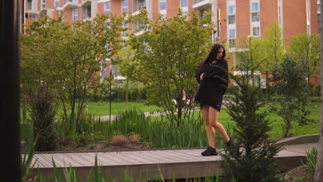 Fashionable-female-with-brown-hair-is-walking-across-the-bridge-looking-around-touching-her-body-wearing-jacket-and-black-trainers-enjoying-time-in-nature-with-beautiful-building-slow-motion