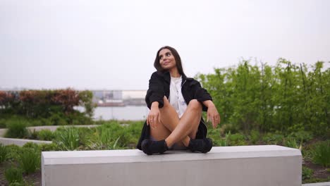 gorgeous-brunette-model-sitting-down-on-grey-bench-in-london-city-park-near-to-a-river-relaxing-posing-wearing-fashionable-trendy-black-jacket-and-trainers-looking-cute-moving-shot-cloudy-weather