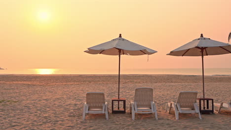 Ein-Paar-Plastiksitze-Und-Sonnenschirme-An-Einem-Leeren-Weißen-Sandstrand-Mit-Goldener-Sonne-über-Dem-Meer-Bei-Sonnenuntergang