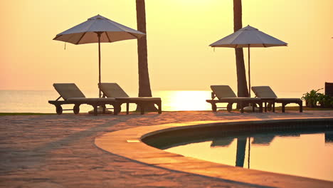 Zwei-Sonnenschirme-Und-Vier-Liegestühle-Am-Pool-Mit-Meer-Im-Hintergrund-Bei-Sonnenuntergang