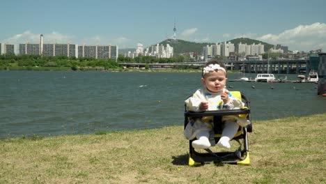 Baby-Girl-Sitting-While-Eating-Outdoor,-Beautiful-River-And-Buildings-In-Background---wide-shot