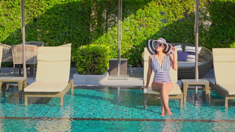 Majestätische-Asiatische-Frau,-Die-Sich-Neben-Dem-Pool-Sonnen-Und-Auf-Einem-Liegestuhl-Im-Royal-Tropical-Resort-Sitzt-Und-Tagsüber-Einen-Abgestreiften-Monokini-Und-Einen-Sonnenhut-Trägt