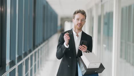 Portrait-of-adult-stylish-man-with-paper-bags-showing-smartphone-and-credit-card-for-shopping
