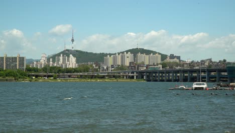 Beautiful-sunny-day-near-Han-River,-Namsan-Tower-and-double-deck-Banpo-bridge-on-background,-catamaran,-and-sailboat-floating-in-the-dock
