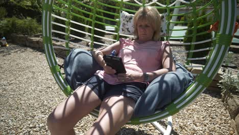 Blonde-woman-in-shorts-reads-eBook-novel-relaxing-in-comfy-garden-hammock-swing-on-hot-sunny-day