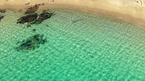 Aerial-pedestal-shot-of-young-woman-walking-into-clear,-calm-emerald-green-water-at-Teurredda-beach-in-South-Sardinia,-Italy-on-sunny-day