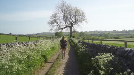 Blonde-woman-with-backpack-on-walking-along-country-track-Ashbourne,-Peak-District,-England