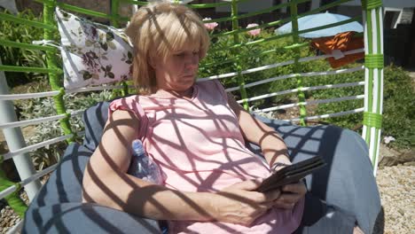Blonde-woman-in-shorts-reads-eBook-novel-relaxing-in-comfy-garden-hammock-on-hot-sunny-day