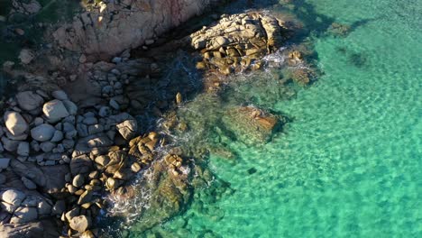 Aerial-shot-of-rocks-in-emerald-green-water-next-to-cliffside-in-South-Sardinia,-Italy