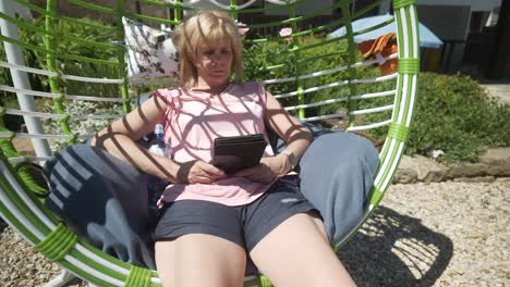 Woman-wearing-shorts-reads-digital-eBook-novel-relaxing-in-comfy-garden-swing-hammock-on-hot-sunny-day