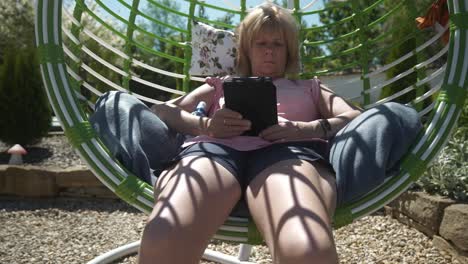 Woman-reading-eBook-novel-relaxing-in-comfortable-garden-swing-hammock-on-hot-sunny-day