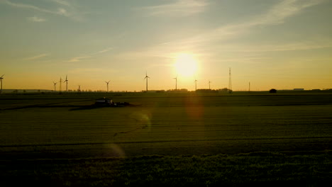 Flying-away-from-the-wind-farm-over-green-fields-on-sunset,-wind-turbines-silhouette-far-away-on-background,-Puck,-Pomorskie,-Poland,-aerial-drone