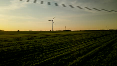 Drone-low-angle-fly-over-green-fields-at-sunset,-wind-turbine-silhouette-on-background