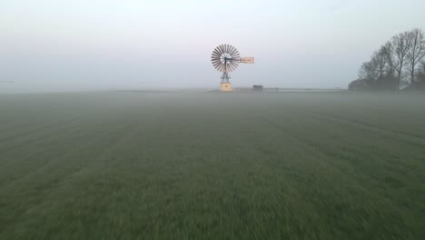 Flying-towards-rotating-steel-windmill-in-misty-farm-field-at-dawn