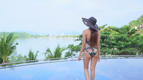 Woman-in-Swimwear-and-Sunhat-Walking-Towards-the-Infinity-Pool-of-Tropical-Island-Resort,-daytime-slow-motion