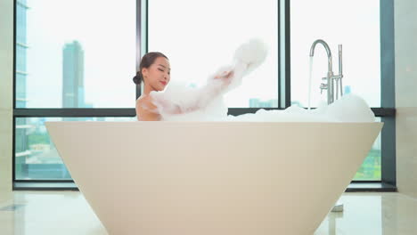 Asian-woman-foaming-her-body,-she-is-lying-in-a-bathtub-full-of-foam-on-a-high-floor-of-the-luxury-apartment-complex-with-glass-walls-in-the-background