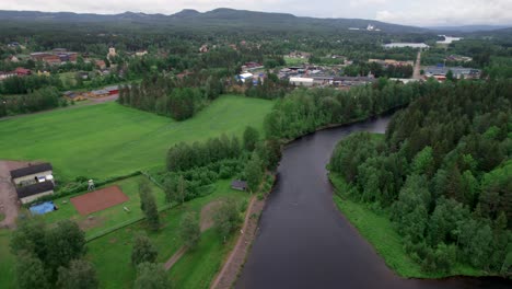 Person-Running-Along-River,-Small-Town-and-Green-Landscape,-Backward-Aerial