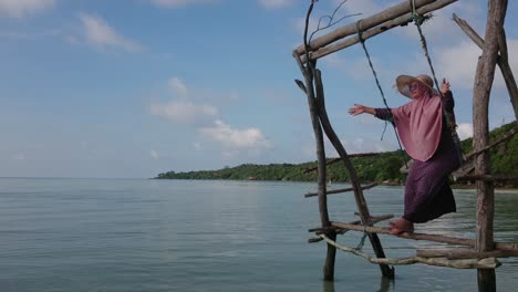 Happy-Woman-in-summer-hat-playing-on-wooden-swing-over-shallow-and-calm-water-at-Karimun-Jawa-island