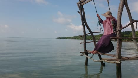Woman-in-summer-hat-swinging-on-wooden-swing-over-shallow-and-calm-water-at-Karimun-Jawa-island-with-camera-tilt-up-and-beautiful-blue-sky-background