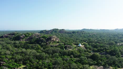 Rocky-outcrop-in-Sri-Lanka-jungle-with-white-Buddhist-shrine,-aerial