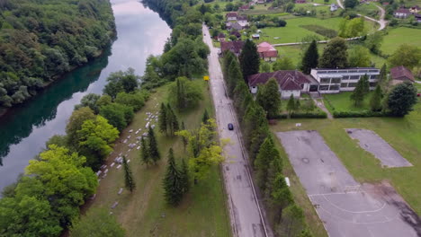 An-aerial-view-of-a-black-car-moving-ahead-with-the-river-and-grave-yard-in-lush-green-town