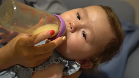 Mom's-hand-holding-the-bottle-with-milk-and-feeding-hungry-baby-holding-baby-in-her-arms,-One-Year-Old-Pretty-Girl-Drinking-Milk-From-Bottle---static-face-close-up