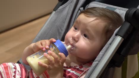 Pretty-baby-girl-drinks-milk-from-bottle-lying-in-a-stroller,-face-close-up
