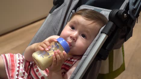 Cute-One-Year-Old-Girl-Drinking-Milk-from-Bottle-Holding-it-with-Both-Hands-while-Lying-in-a-Stroller-and-Looking-Around---close-up,-static-shot