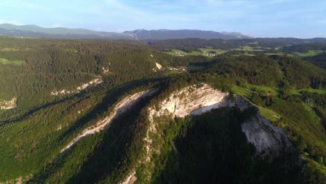 Aerial-flyover-overgrown-mountain-of-Belvedere-de-la-Roche-Blanche-and-in-background-Monts-Jura-during-spring-season-and-sunlight