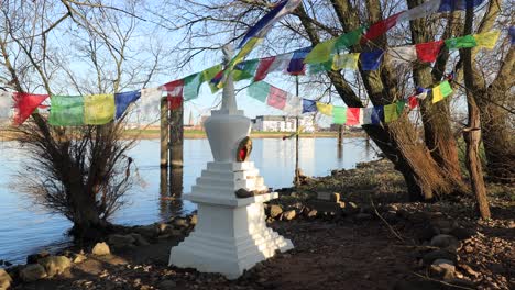 Intime-Szene-Eines-Kleinen-Buddhistischen-Stupa-Ornaments-Am-Rande-Des-Flusses-Ijssel-In-Zutphen,-Umgeben-Von-Bäumen-Und-Vegetation-Mit-Bunten-Gebetsfahnen,-Die-Im-Wind-Wehen