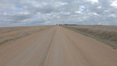 Point-of-view-driving-on-a-straight-stretch-of-gravel-road-and-past-a-farm-yard-with-grain-bins-and-barn