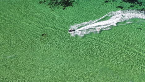 Aerial-view,-jetski-watercraft-towing-skier-in-clear-shallow-ocean-water-of-west-Australia,-top-down-tracking-drone-shot