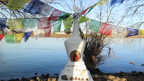 Nahaufnahme-Eines-Kleinen-Buddhistischen-Stupa-Ornaments-Am-Rande-Des-Flusses-Ijssel-In-Zutphen,-Umgeben-Von-Bäumen-Und-Vegetation-Mit-Bunten-Fahnen,-Die-Sanft-Im-Wind-Wehen,-Und-Wasser,-Das-Sich-Im-Rücken-Kräuselt