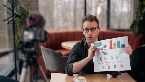 Stylish-young-man-with-glasses-makes-a-presentation-on-slides-infographics-and-records-video-on-camera-on-a-tripod