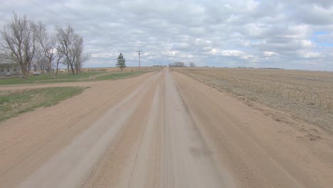 Point-of-view-driving-on-a-straight-stretch-of-gravel-road,-past-a-farm-yard,-a-crossroad-and-harvested-fields