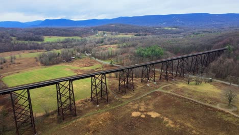 Aerial-drone-video-footage-of-a-train-bridge-viaduct-running-over-a-valley-in-the-Appalachain-Mountains-during-early-spring-on-a-cloud-day,-surrounded-by-mountains-and-farmland